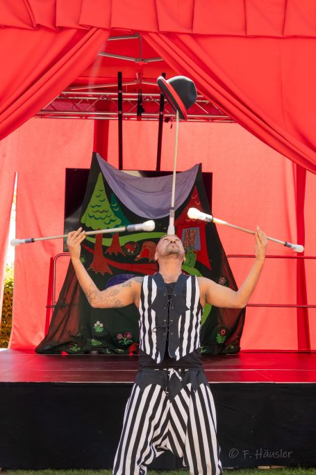 Jongleur balanciert einen Hut auf seinem Kopf und hält Stäbe in den Händen.