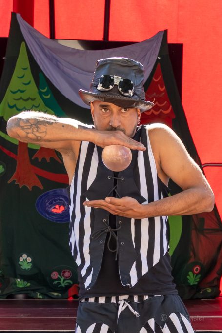 Ein männlicher Performer mit Hut und gestreiftem Outfit zeigt eine Jonglage mit einer Kugel.