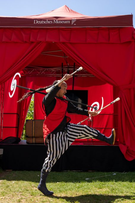 Jongleur in gestreifter Hose tanzt vor einem roten Zelt und jongliert mit Stöcken.